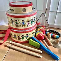 Photo showing two vintage children's drums, maracas, wooden clapper, mini cymbals, drumsticks, and two tambourines on wooden floor