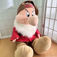 Front view of large vintage Disney plush Grumpy seated, showing full body with red jacket, brown hat embroidered with "GRUMPY," and white faux fur collar.