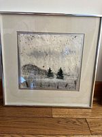 Framed print showing two small trees on a textured, muted background with some spotting, behind glass in a simple metal frame.