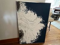 Frontal view of the framed wave abstract painting leaning against a wall on wood floor.