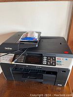 Front and top view of Brother multi-function printer with control panel and screen, showing some paper and cables on top.