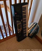Yamaha electric keyboard, black, 49 keys, with digital controls and display, placed vertically on carpet floor near railing and mirror.
