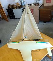 Model sailboat standing upright on a table, showing large white sail with numbers and logo, hull design with blue and green accents.