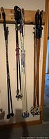 Photo of eight ski poles hanging on a wooden wall rack, including a mix of downhill and cross country styles, showing handles, baskets and various materials