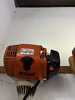 Close-up photo of an orange Stihl lawn tool engine, showing wear and dirt, with a barcode sticker on top.