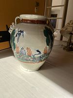 Front angled view of the ceramic vase on a white table, clearly showing painted birds, plants, the brown band, and loop handles.