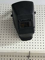 A black plastic welders mask hanging on a pegboard with a flip-up lens window.