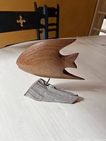 Side view of the medium-brown wooden fish sculpture on driftwood base, showing smooth carving with stylized fins and tail, placed on table with black chairs in background.