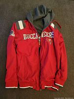 Full front view of the red side of the reversible Buccaneers jacket with gray hood inside and Buccaneers logos visible.