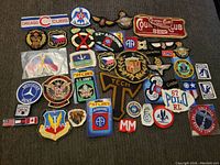 Collection of various embroidered patches laid out on a dark surface showing military, automotive, and vintage advertising designs.
