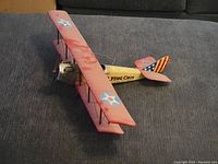 Front-side view of a vintage model biplane airplane with red wings marked with blue and white star insignia, yellow fuselage, and American flag design on the tail, placed on a gray surface.