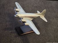 Silver vintage motor airplane model with two engines and propellers, one propeller missing, mounted on a black rectangular stand, on carpeted floor.