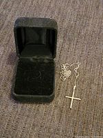 Black velvet jewelry box open with 14k gold cross pendant and chain placed beside it on fabric surface.