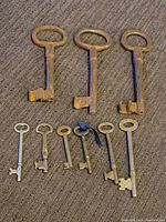 Ten skeleton keys laid out on a fabric surface, three large rusty keys on top and seven smaller silver-colored keys below.