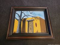 Framed painting showing an adobe house with bare tree and blue sky, with dimensions indicated by dollar bill for scale.