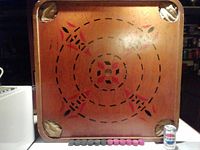 Photo of crokinole side of the wooden game board showing scoring circles, red and black painted areas, and four fabric corner pockets. Pieces shown below the board for scale.