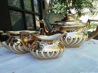 Tea set including teapot, sugar bowl, and creamer seen on a white cloth with floral tablecloth underneath