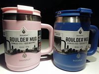 Front view of two MANNA Boulder Mugs, one pink and one blue, showing the labels and stainless steel body with city skyline graphics.