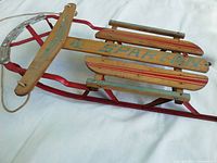 Photo angled showing the front and side of the mid-century Spartan sled with red metal frame and wooden slats showing blue Spartan text.