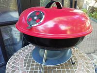 Side view of the red lid and black base tabletop charcoal barbecue on a tiled outdoor table.