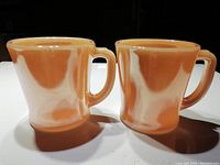 Two peach lustre Fire King mugs shown side by side, highlighting color and handle shape.