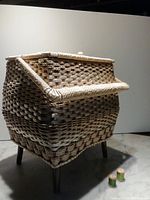 Woven wicker sewing basket with hinged removable lid resting on black wooden legs, close-up view of basket side and lid detail.