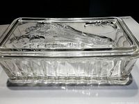 Side view of glass refrigerator dish with lid showing embossed vegetable designs including celery, tomato, and radishes.