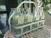 Front angle of metal carrier filled with eight clear glass milk bottles on table outdoors.