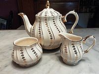 Full view of teapot, milk creamer, and sugar bowl with gold swirl decoration on white ceramic on marble countertop.