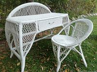 Full view of white wicker vanity makeup table and matching chair placed outdoors on grass with foliage in background.