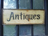 Front view of wooden antiques sign showing weathered light wood background with black text 'Antiques' and darker wood frame.