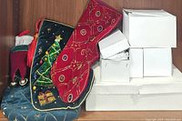 Photo showing four Christmas stockings including blue velvet with tree motif, red star patterned, personalized red with white cuff, and blue with bells alongside four white boxes containing ornaments.