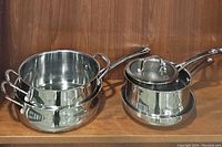 Four stainless steel cooking pots grouped together, including two-handled larger pots and saucepans with lids.