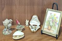 Small framed floral painting, porcelain gnome figurines, clam shell shaped jewelry case, and porcelain clam shell box on wooden surface.