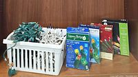 Photo showing white basket containing multiple loose strands of holiday string lights in green and white wires, and five boxed sets of holiday lights arranged in a row behind it.