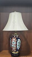 Full view of Asian porcelain jar lamp showing traditional painted scenes and cream fabric shade.