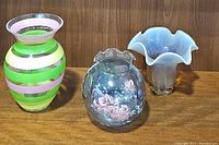 Three vases displayed side by side on a wooden surface against a wood panel background. Pink & green striped vase on left, round floral painted vase in middle, open flower shaped vase on right.