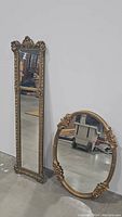 Photo showing both mirrors leaning against a wall on a concrete floor. Rectangular mirror is tall and slender with detailed floral and scrollwork gold frame. Oval mirror features raised floral decorations on gold frame.