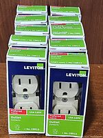 Photo showing 9 boxes of Leviton ivory electrical outlets, model CBR15, 15A 125V, Preferred series, stacked on a wooden surface.