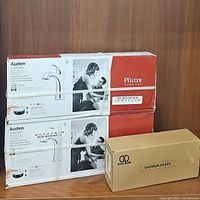 Two boxed Pfister Auden faucets and a boxed Doliese showerhead on wooden surface with brown background