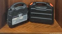 Two black molded plastic cases side by side on wooden surface. One labeled Craftsman with visible translucent compartment, the other plain with orange latches.