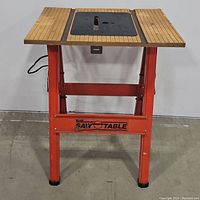 Front view of vintage portable saw table showing wooden slatted surface with central black metal insert and orange legs.
