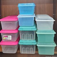 Stack of assorted storage boxes with translucent bases and colored lids in pink, turquoise, and blue shown stacked inside a wooden cabinet.