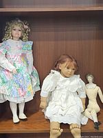Three dolls displayed together on a wooden shelf: Annette Himstedt doll, floral dressed curly hair doll, and antique porcelain head doll