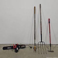 Full view of electric chain saw and three yard tools against a white wall showing complete lot contents