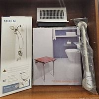 Lot showing Moen showerhead in box, folding shower seat box, wrapped Moen grab bar, and two white air vents stacked on top of each other