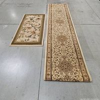 Two rugs side by side on a floor; a long ornate runner rug on the right with tan and brown floral motifs and a smaller floral rectangular rug on the left.
