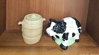 Two cookie jars displayed on wooden shelf; left is beige barrel shaped Frankoma pottery jar, right is black and white Holstein cow ceramic cookie jar.