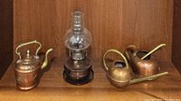 Photo showing the copper tea kettle, copper oil lamp with glass chimney, and two small copper watering cans on a wooden shelf.