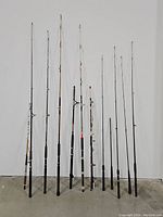 Full view of 12 fishing rods of varying lengths and handles against white wall and concrete floor.